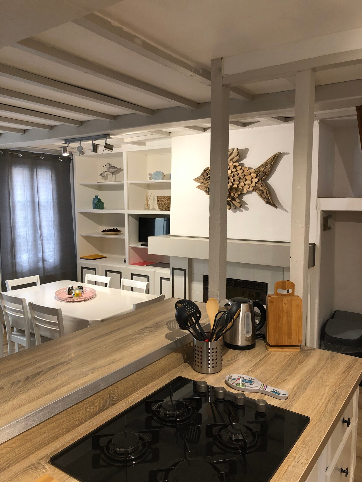 The kitchen area is equipped with modern appliances and utensils, featuring a gas cooktop and a wooden cutting board. A dining table is visible in the background, surrounded by white chairs. Decorative shelves display various items, including a wooden fish sculpture, creating a functional yet stylish space.