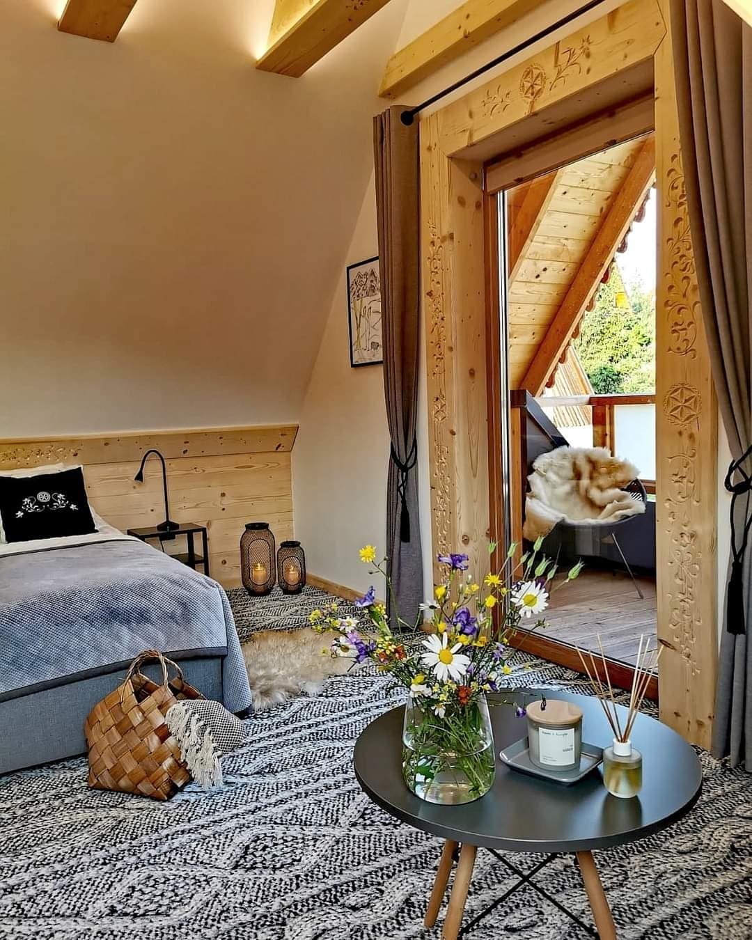 A bedroom features wooden beams and a cozy atmosphere, with a spacious bed dressed in gray linens. A small table holds a vase of fresh flowers and a candle. A balcony door opens to a scenic view, with a comfortable chair and decorative throw.