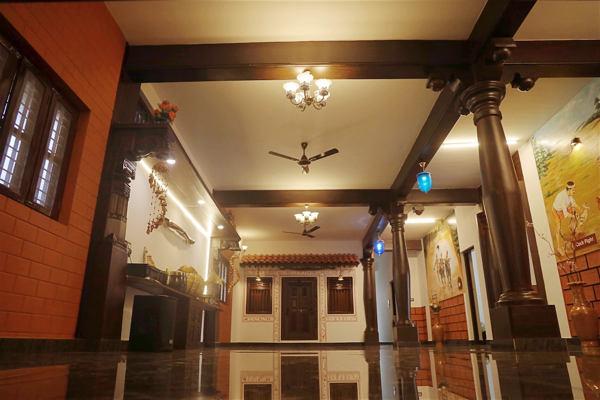 A spacious room is highlighted by decorative wooden pillars and ceiling fans. Traditional art pieces adorn the walls, complemented by subtle lighting. The polished floor reflects the vibrant colors of lanterns hanging from the ceiling, creating a welcoming ambiance.