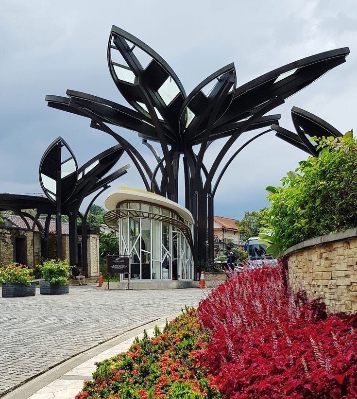 An eye-catching entrance structure features modern black leaf-like architecture, accompanied by a gracefully curved glass door. Vibrant red and orange flowers line the pathway, adding a splash of color against the natural stone and greenery in the surrounding area.