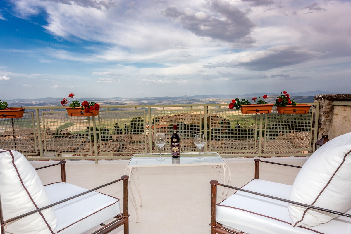 A spacious terrace is showcased with two comfortable armchairs positioned around a glass table. Potted red flowers adorn the railing, overlooking a panoramic view of the rolling hills of the Tuscan countryside under a partly cloudy sky.