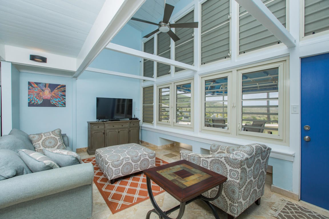 A bright living area features large, floor-to-ceiling windows, allowing natural light to fill the space. A soft sectional sofa and matching ottoman provide comfortable seating, complemented by two armchairs. A flat-screen television rests on a dark wooden console, adding to the room's functionality.