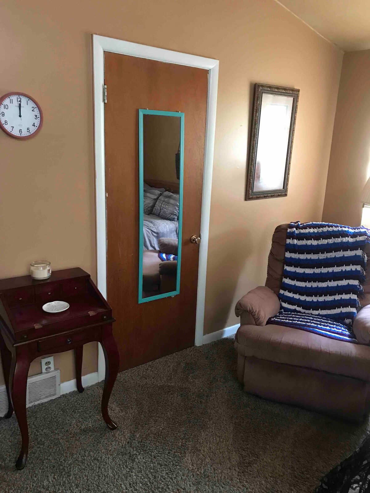 A cozy corner of a room is depicted, featuring a wooden side table with a plate and candle atop. A comfortable armchair is present, positioned near a door with a mirror, and light is softly reflected on the wall. A framed mirror hangs above the chair, enhancing the area.