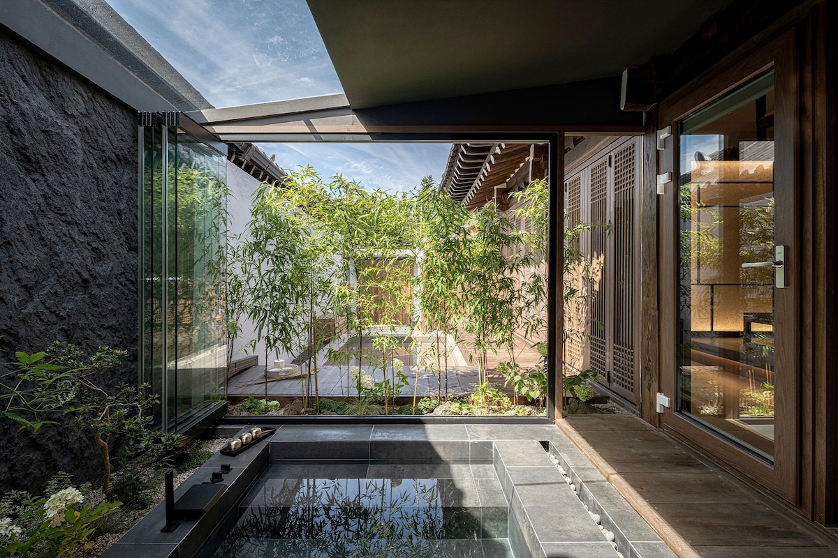 An indoor space features a sleek, modern jacuzzi surrounded by glass walls, allowing views of a serene garden filled with green plants. Natural light floods the area, enhancing the connection between indoor luxury and outdoor tranquility.
