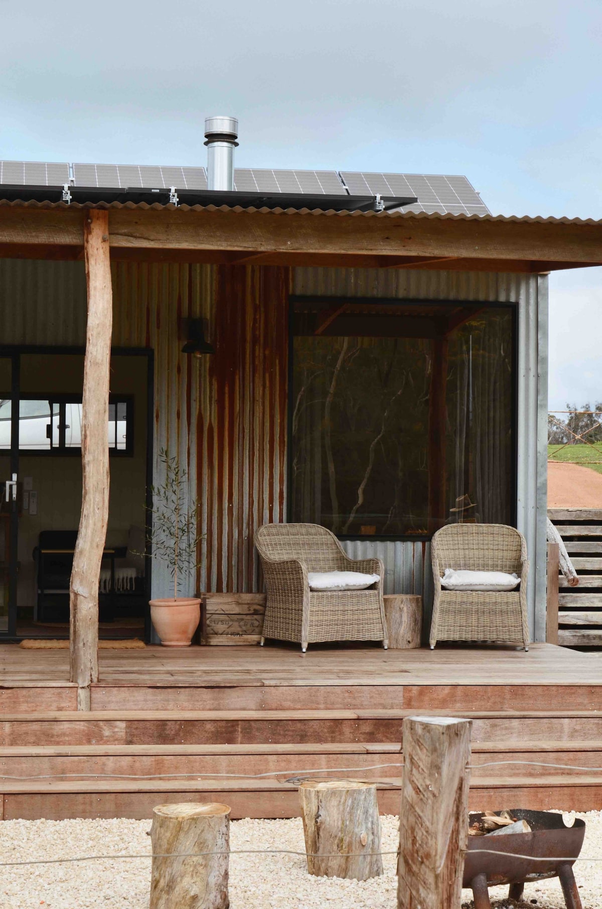 A rustic exterior of Cleves Hut features a deck with two woven chairs, inviting relaxation. The porch is framed by wooden supports, with a window and doorway providing access to the interior. Solar panels are visible on the roof, emphasizing an eco-friendly design.