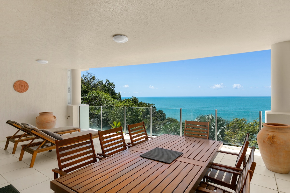 A spacious balcony is furnished with a wooden dining table and chairs, and sun loungers, providing a serene space to enjoy the stunning ocean view. Lush greenery is visible at the edges, with the sparkling Coral Sea stretching beyond under a clear blue sky.