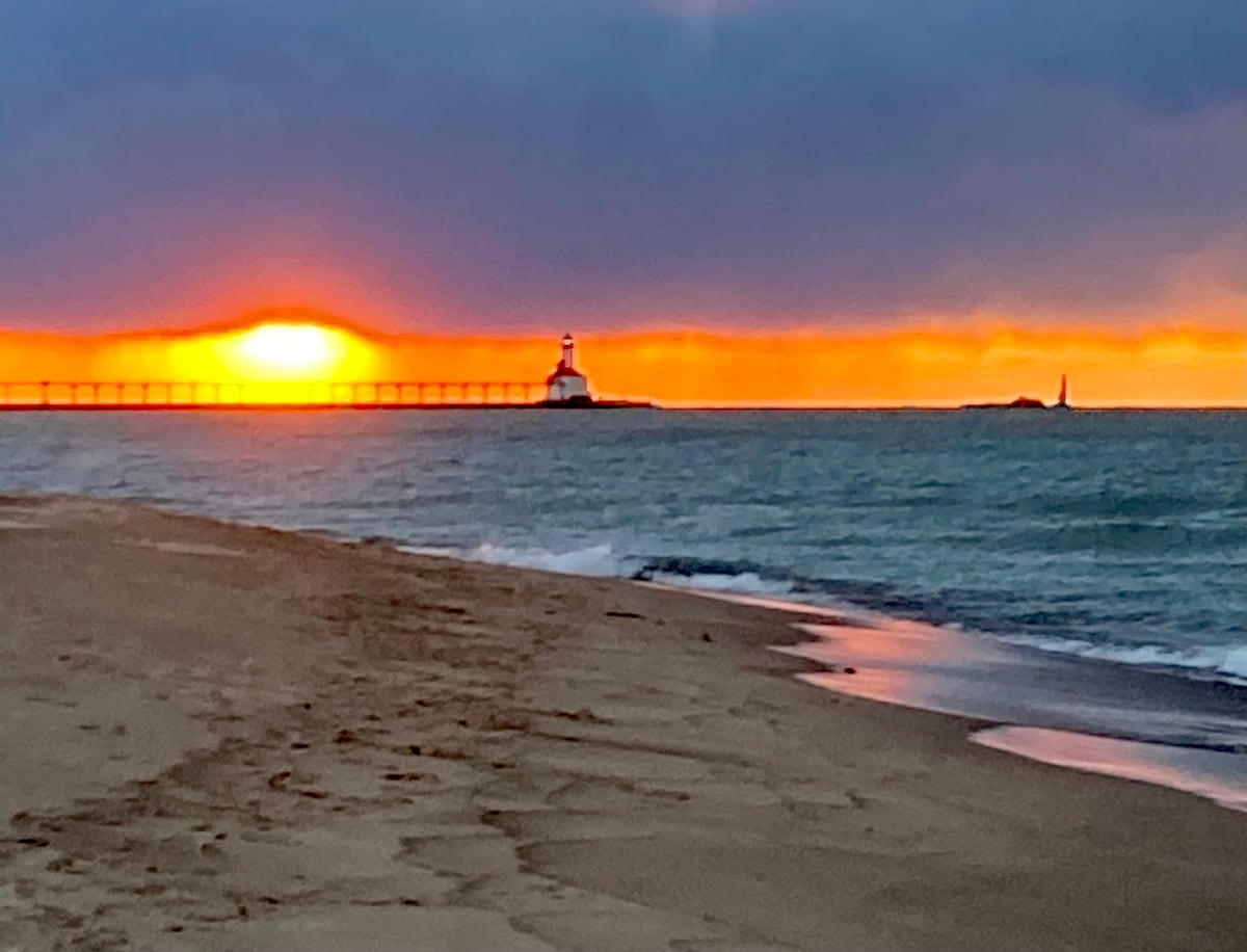 A serene lake view captures the sun setting over the horizon, casting warm orange and yellow hues across the sky. Gentle waves lap against the sandy beach, while a lighthouse stands in the distance, framed by the soft glow of twilight.