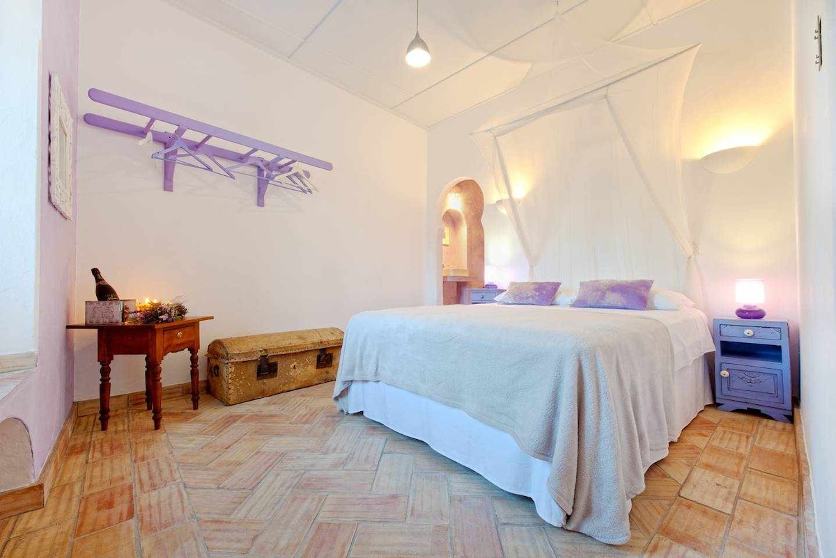 A spacious room is presented with soft lilac accents. A double bed, draped in a light-colored cover, is featured prominently. A wooden table sits beside the bed, accompanied by a decorative trunk. A wall-mounted coat rack adds a touch of functionality to the space.