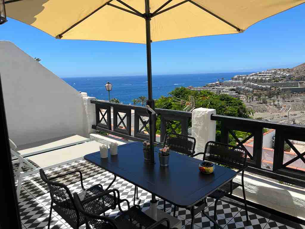 A spacious terrace is shown, featuring a black dining table surrounded by four chairs. Two lounge chairs are positioned beside the railing. An umbrella provides shade while the ocean view extends towards the horizon, with the vibrant landscape of nearby hills visible in the background.