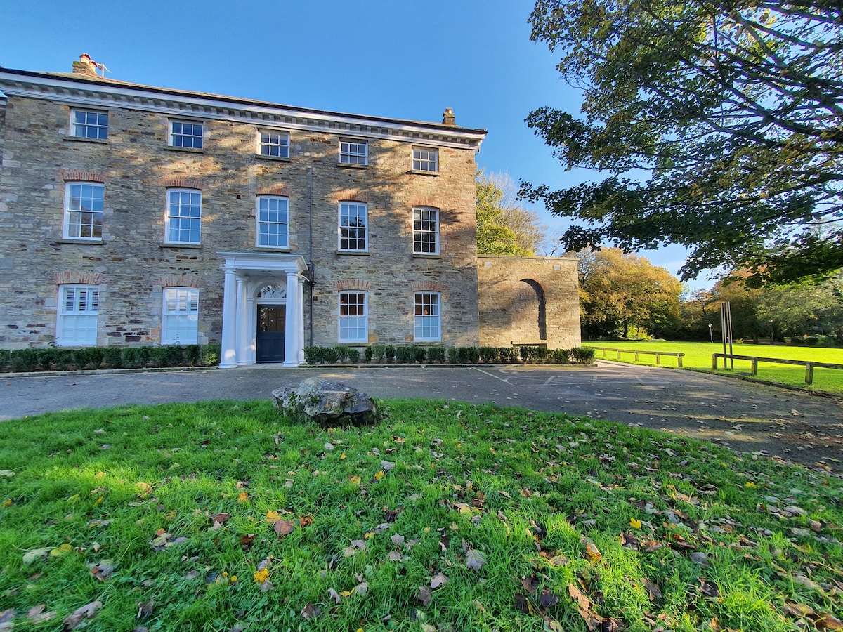 The historic building features a stone facade with large windows, set against a clear blue sky. A well-maintained lawn with scattered fallen leaves leads to the entrance, framed by trees and a grassy area, offering a serene view of the surrounding landscape.