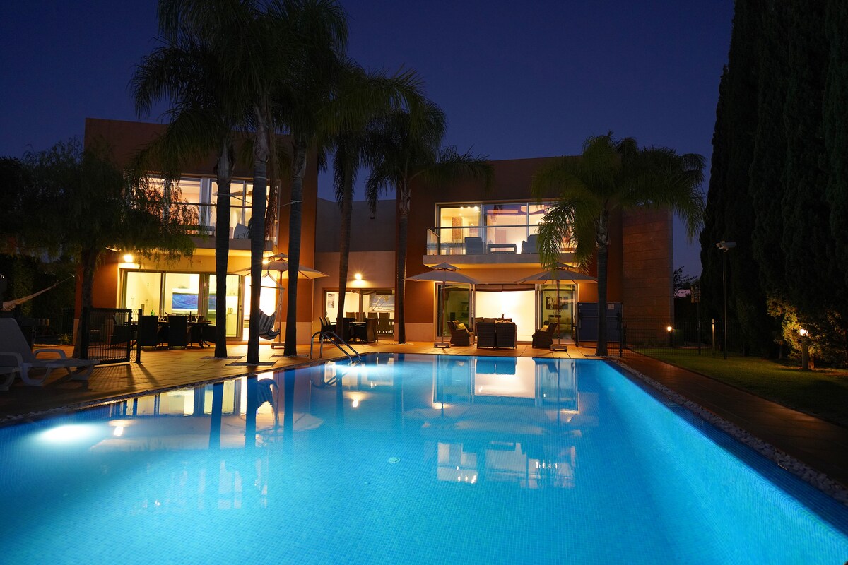 The villa is showcased at twilight, with large glass windows reflecting warm indoor lighting. A serene pool spans the foreground, its surface illuminated in vibrant blue. Lush palm trees frame the scene, creating an inviting outdoor space for relaxation.