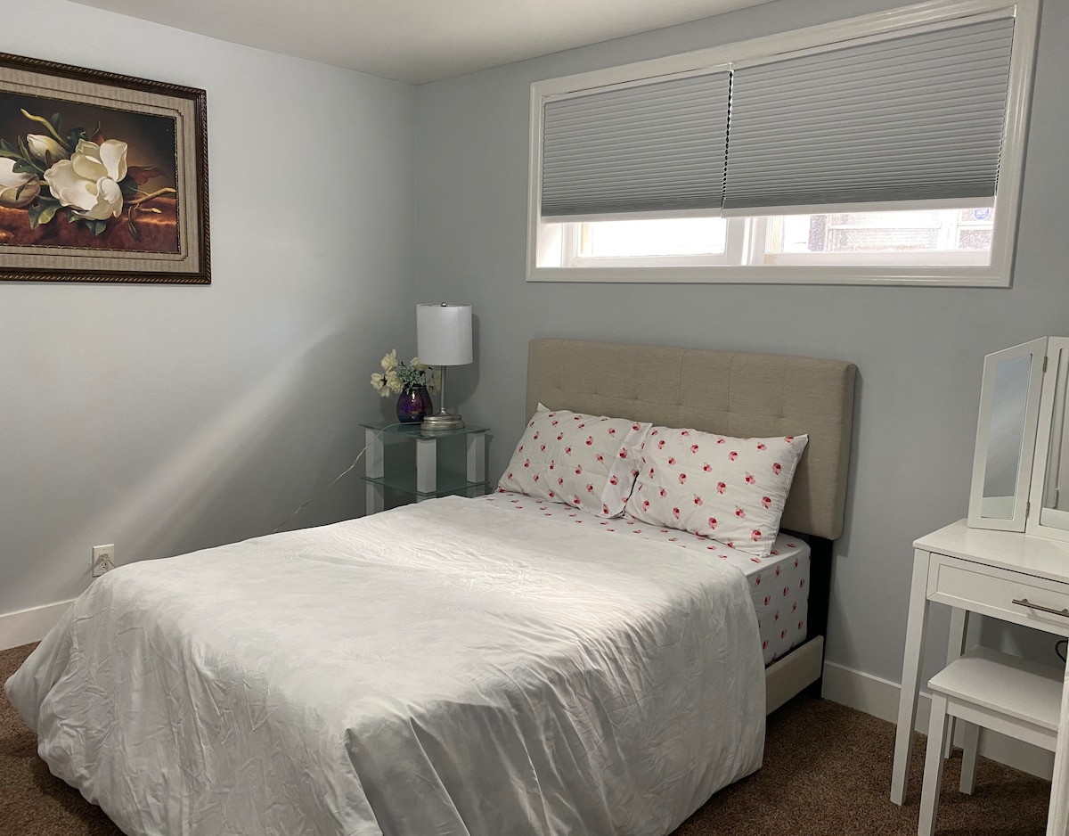 A well-lit sleeping area features a spacious bed with a light-colored quilt and decorative pillows. A framed floral artwork adorns the wall. A bedside lamp and a small floral arrangement are placed on a glass table, while a dresser with a mirror completes the room's setup.