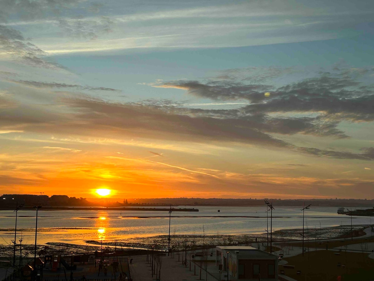 A sunset casts warm hues over the river, reflecting beautifully on the water’s surface. Silhouettes of nearby trees and lampposts create a serene atmosphere, while the horizon showcases the gentle curve of the landscape in the distance.