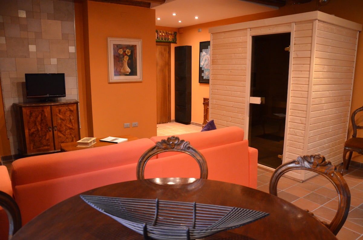 A cozy living space features an orange sofa and a wooden dining table adorned with a decorative centerpiece. A sauna is visible in the corner, while a flat-screen TV is mounted on the wall. Soft lighting enhances the warm atmosphere of the room.