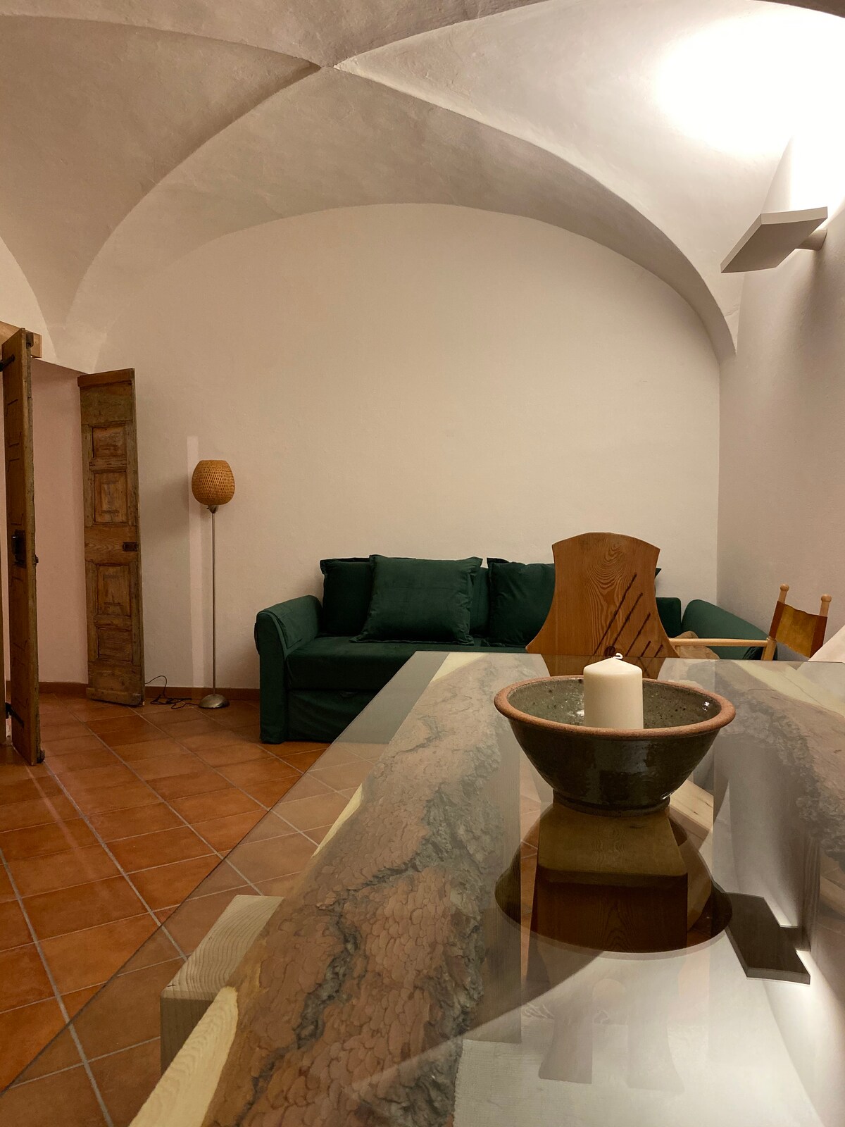 A cozy living area is visible, featuring a green sofa and wooden dining chairs around a marble-top table. Warm light is provided by a standing lamp, highlighting the arched ceiling and terracotta floor tiles.