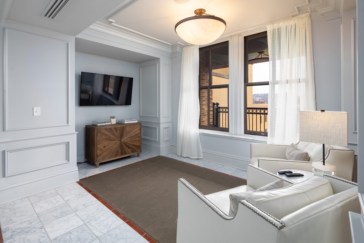 A spacious living area is enhanced by natural light from large windows with sheer white curtains. A comfortable white armchair and sofa are positioned around a neutral-toned area rug. A wooden media console holds a flat-screen TV, while sleek marble floors complement the classic architectural details.