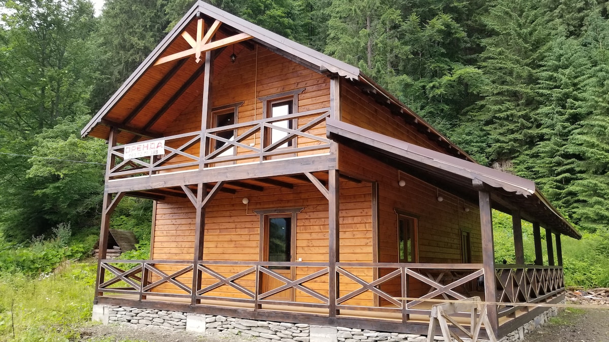 A wooden chalet is surrounded by lush greenery, featuring a spacious porch with wooden railings. The two-story structure presents large windows and a steeply pitched roof, blending harmoniously with the forested landscape.