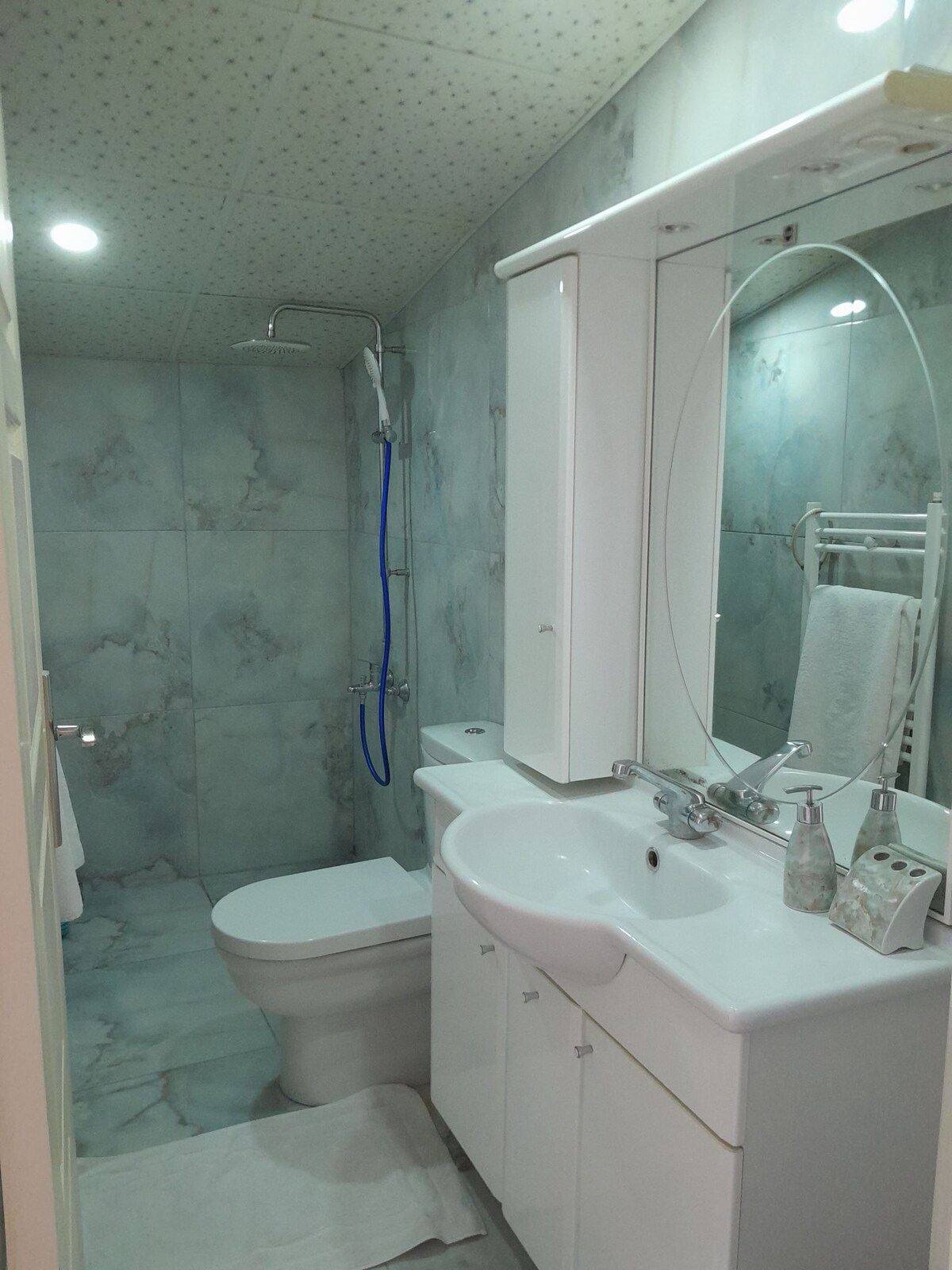The bathroom features modern fixtures and light-colored, marbled walls. A walk-in shower is fitted with a clear glass door and a chrome showerhead. The spacious vanity includes a circular mirror, a sink with decorative containers, and ample storage beneath.