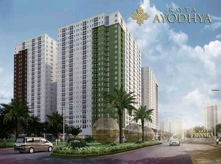 A modern apartment complex is shown with a blend of white and green facades, surrounded by landscaped greenery. The building features numerous windows and balconies, providing a view of the street lined with palm trees and well-maintained roads.