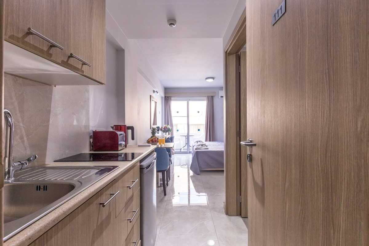 A modern kitchenette is visible on the left, featuring a sink and essential appliances. The open layout leads to a well-lit living area with a dining table and chairs. A spacious bed in the distance is set against a large window, allowing natural light to fill the room.