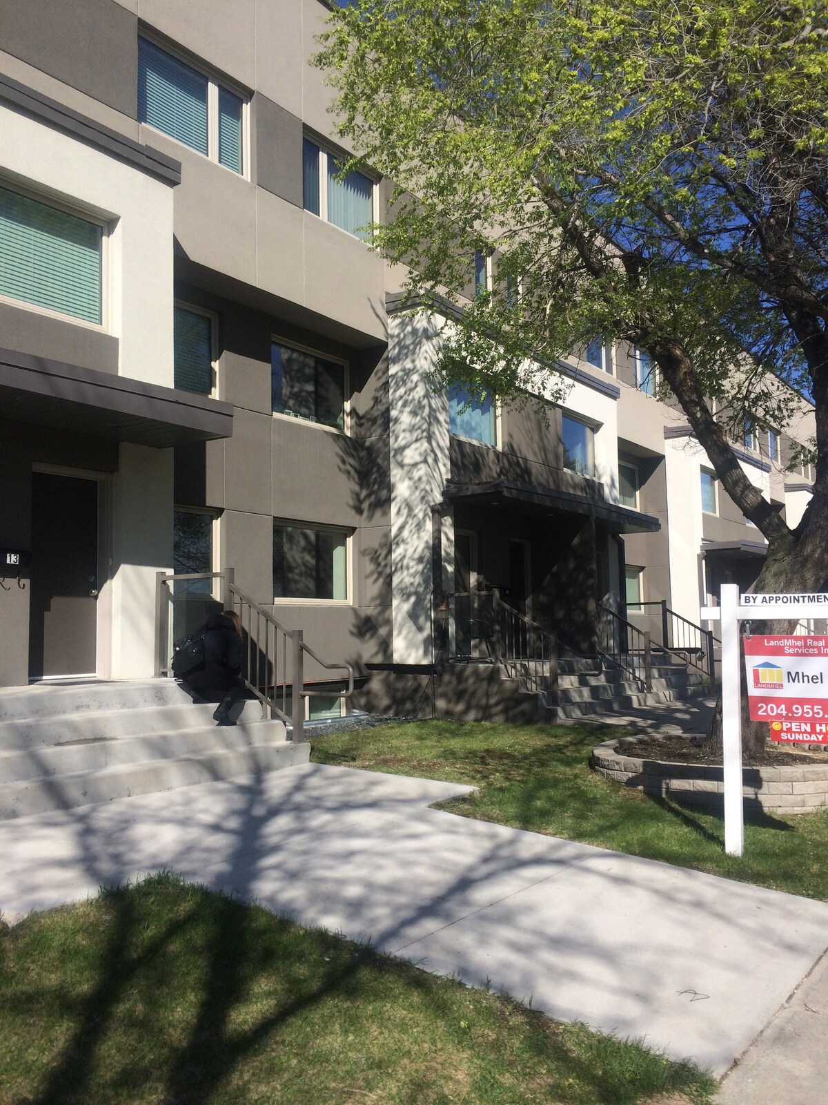 The exterior of a modern multi-story building is shown, characterized by geometric designs and large windows. Steps lead up to the entrance, alongside a manicured lawn with trees casting shadows on the pathway. A for sale sign is also visible in the foreground.
