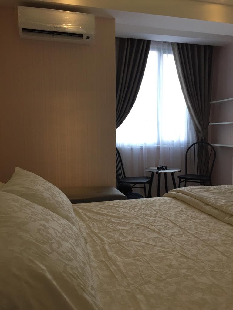 A soft-colored bedroom features a spacious bed with light bedding, creating a calm ambiance. A window draped with sheer curtains allows natural light to enter. Two black chairs and a small table are positioned beside the window, offering a cozy seating area.