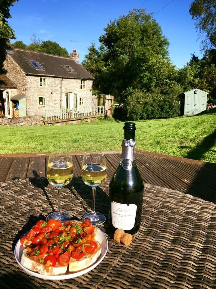 A table set on a wooden terrace features two glasses of white wine beside a plate of vibrant tomato bruschetta. In the background, the stone cottage is framed by lush greenery under a clear blue sky, enhancing the natural setting.