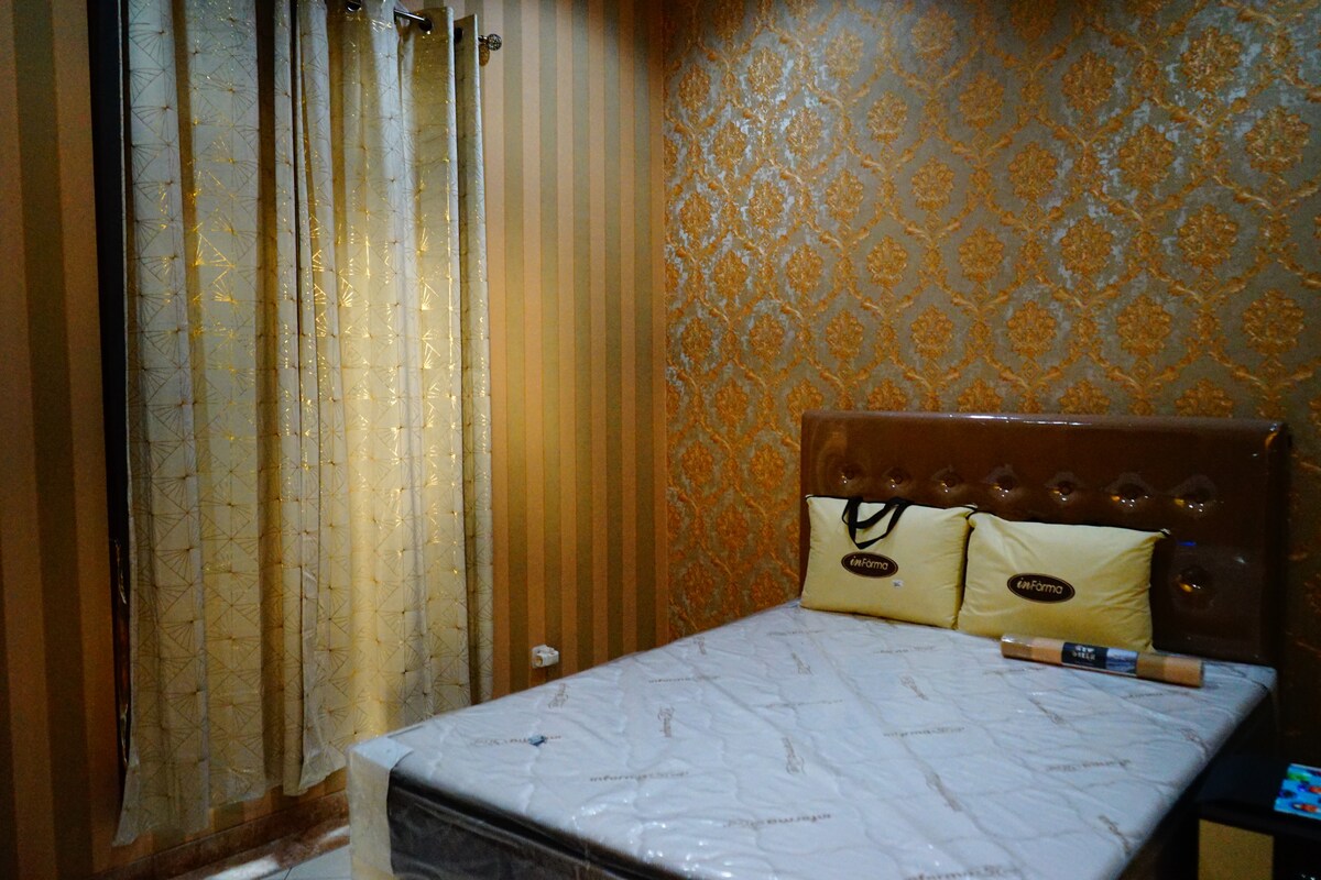 A comfortable sleeping area is noted, featuring a spacious bed with decorative pillows. The walls are accented with elegant gold patterns, while striped drapes cover a window, allowing soft light to enter the room.