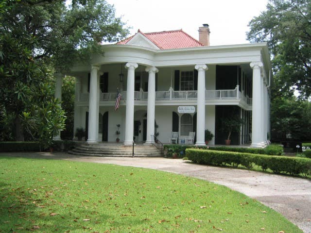 A historic home is showcased with a classic architectural style and a prominent red-tiled roof. White columns support the large front porch, which features rocking chairs. Manicured lawns and trimmed hedges frame the entrance, adding to the overall charm and inviting appearance.