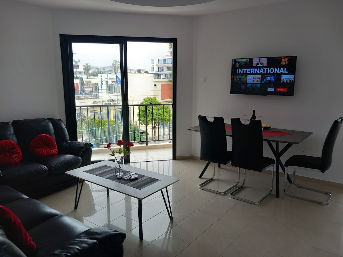 A modern living space is highlighted by a large window that offers natural light and views of the outdoor area. A comfortable black sofa with red accent pillows is paired with a sleek coffee table. A dining table for four is seen adjacent to a wall-mounted television.