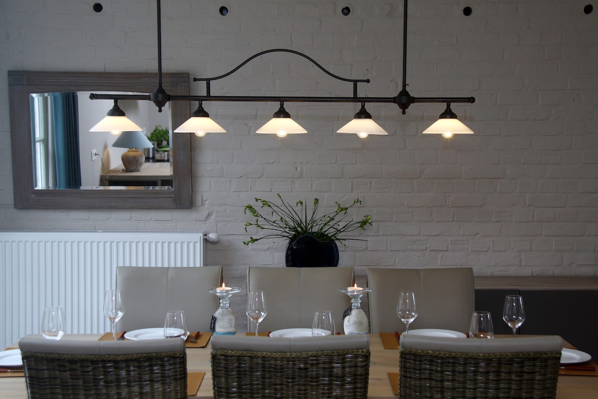A spacious dining area is highlighted by a long wooden table set with plates, glassware, and candles. Five pendant lights illuminate the table, while a decorative mirror and a vase with greenery adorn the walls, creating an inviting meal setting.
