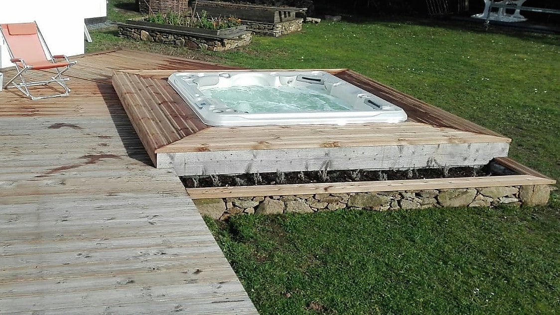 A wooden terrace is featured, surrounding a hot tub set on a raised platform. Lush green grass is present, and a single lounge chair is positioned nearby on the deck, creating a relaxing outdoor space.