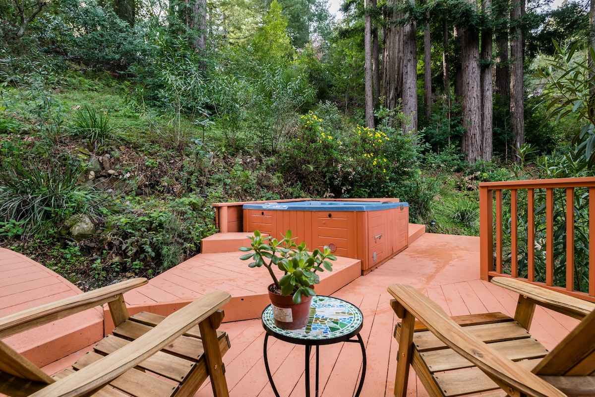 An outdoor deck is surrounded by lush greenery and towering redwoods. A hot tub is situated on a raised platform, while two wooden Adirondack chairs face a small side table featuring a potted plant, creating a serene space for relaxation.