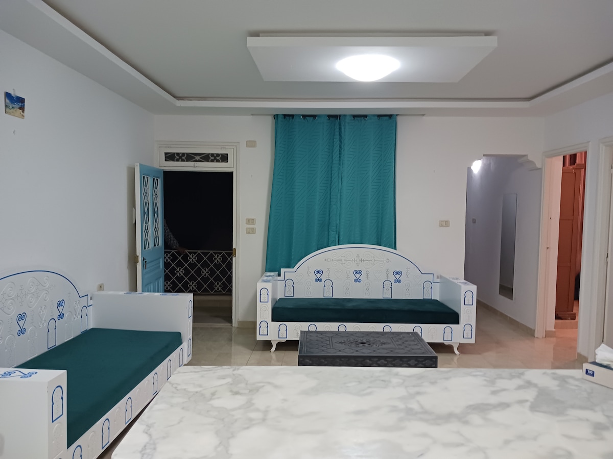 A light-filled living area features two upholstered sofas with turquoise cushions, complementing turquoise drapes that frame a spacious window. A marble-topped coffee table is positioned centrally, and the area flows into a hallway that leads to additional rooms.