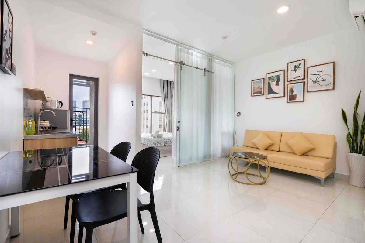 A bright and airy living area is depicted with a light-colored sofa and decorative cushions. A glass-top coffee table is positioned in front, and a dining area with black chairs is visible. Large glass doors offer views of the city skyline and allow natural light to fill the space.