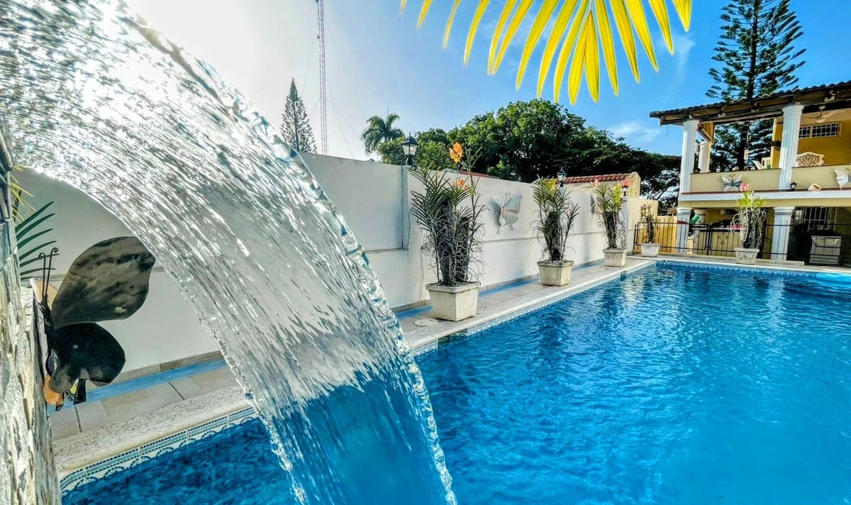 A clear view of a private swimming pool features a gentle waterfall cascading into the water. Lush greenery and decorative plants frame the pool area, while a spacious deck and seating arrangements are visible in the background, promoting relaxation.