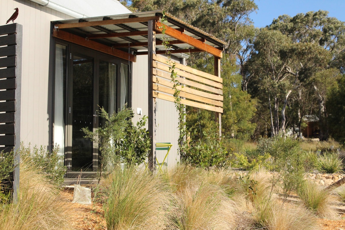 The exterior of the studio features a wooden awning supported by beams, with large glass doors providing a view into the living space. Native shrubs and grasses surround the building, blending it with the natural landscape and offering a serene outdoor environment.