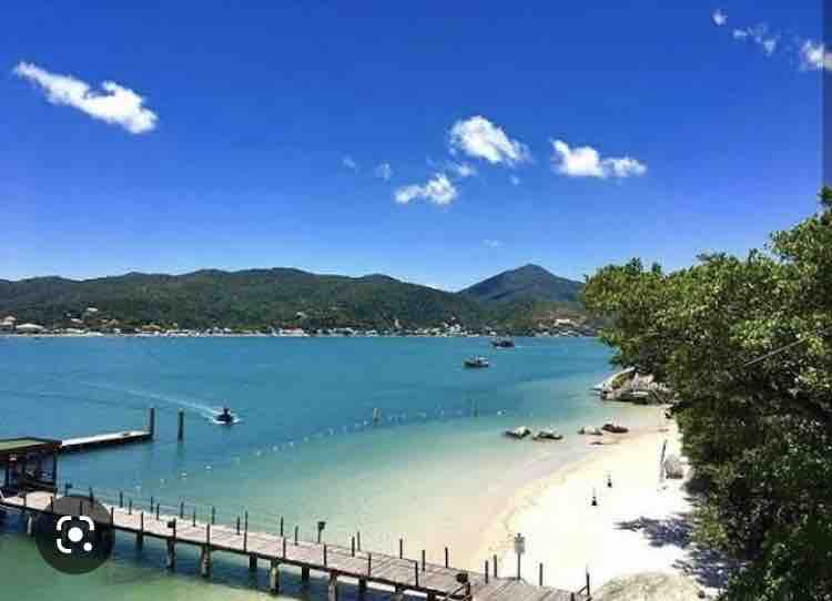 A scenic waterfront view captures the blue waters of the bay under a clear sky dotted with a few clouds. A wooden pier extends into the water, accompanied by small boats and a beach with soft, light sand. Lush greenery frames the scene.