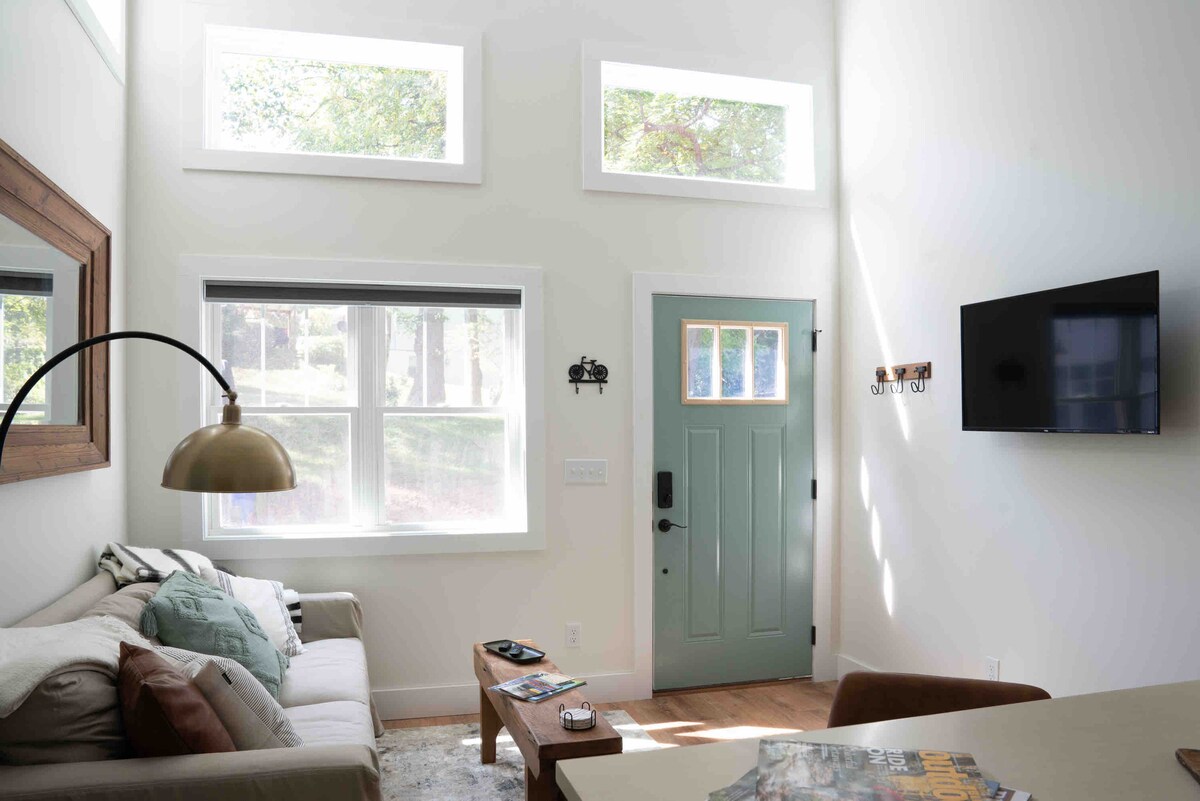 A bright living space is presented with high vaulted ceilings and large windows that allow natural light to fill the room. A comfortable sofa, a wooden coffee table, and a wall-mounted TV are visible. The exterior door features a refreshing light green color.