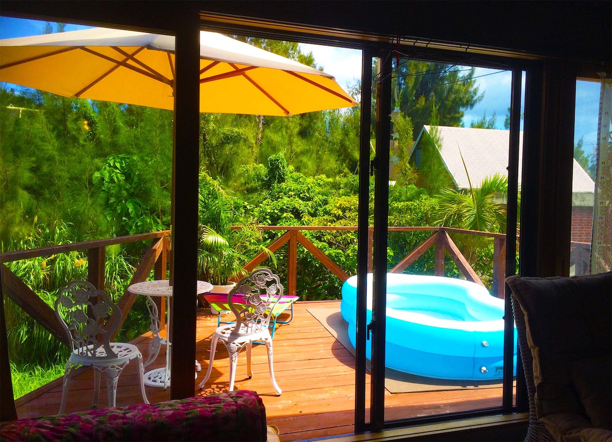 A private deck is revealed through large glass doors, adorned with a blue jacuzzi and two white chairs beside a small table. A yellow umbrella provides shade, while lush greenery frames the peaceful outdoor space.
