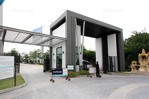 The entrance features a modern architectural design with tall, sleek structures framing an open passage. Lush greenery is present on either side, and a guard can be seen monitoring access. The paved driveway leads into a serene community environment.