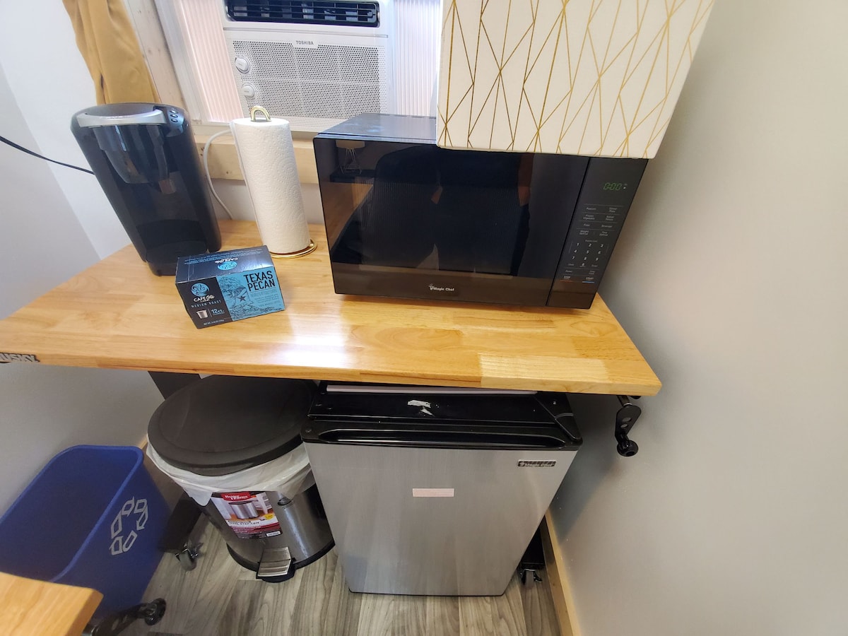 A modern kitchen area features a wooden countertop, equipped with a microwave and coffee maker. Below, a small refrigerator is placed next to a recycling bin, and a roll of paper towels is nearby, creating a functional and organized workspace.