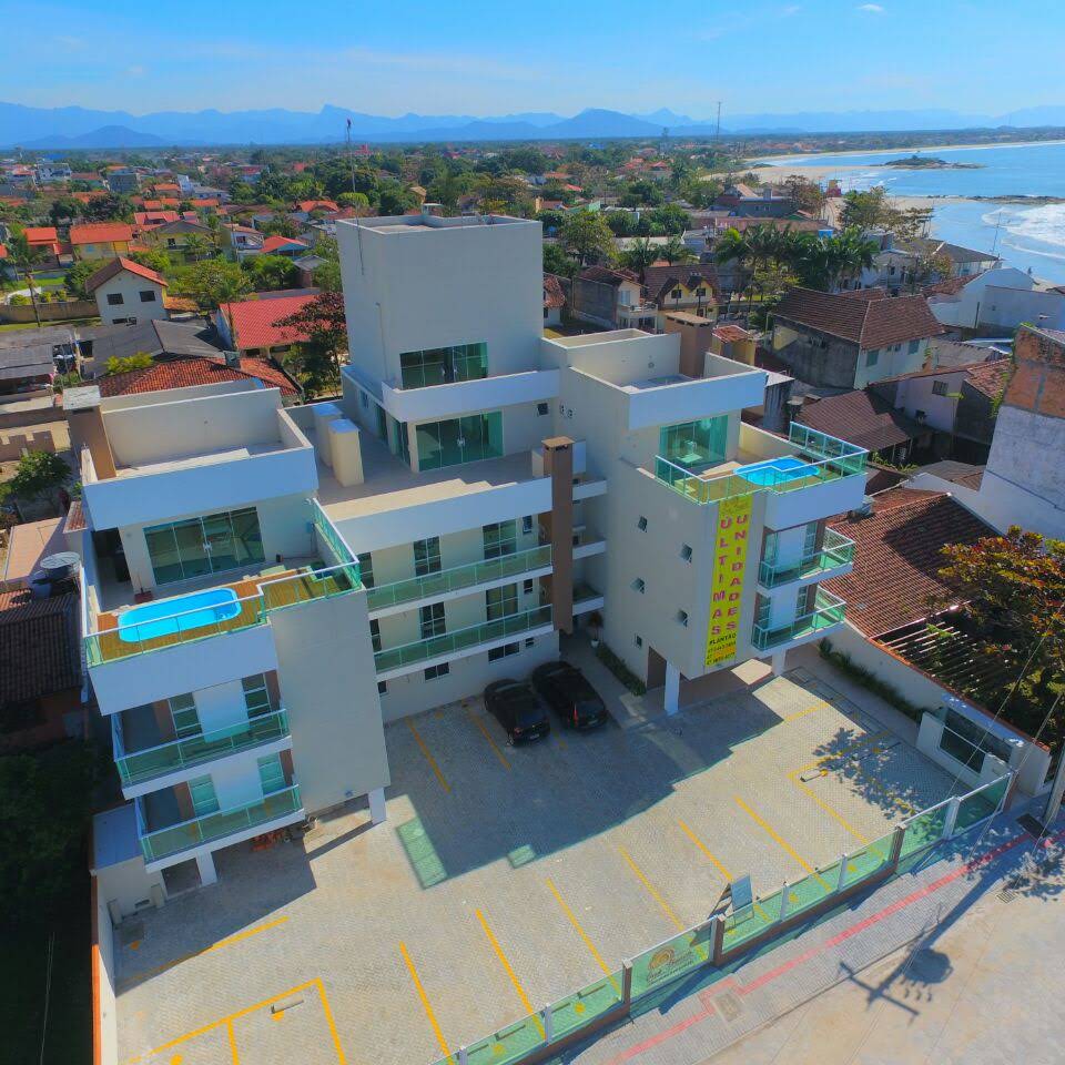 An aerial view captures a modern multi-story building with a light-colored façade and balconies. The surrounding area features a parking lot with designated spaces. In the background, the coastline and peaceful ocean waves are visible, framed by greenery and residential structures.