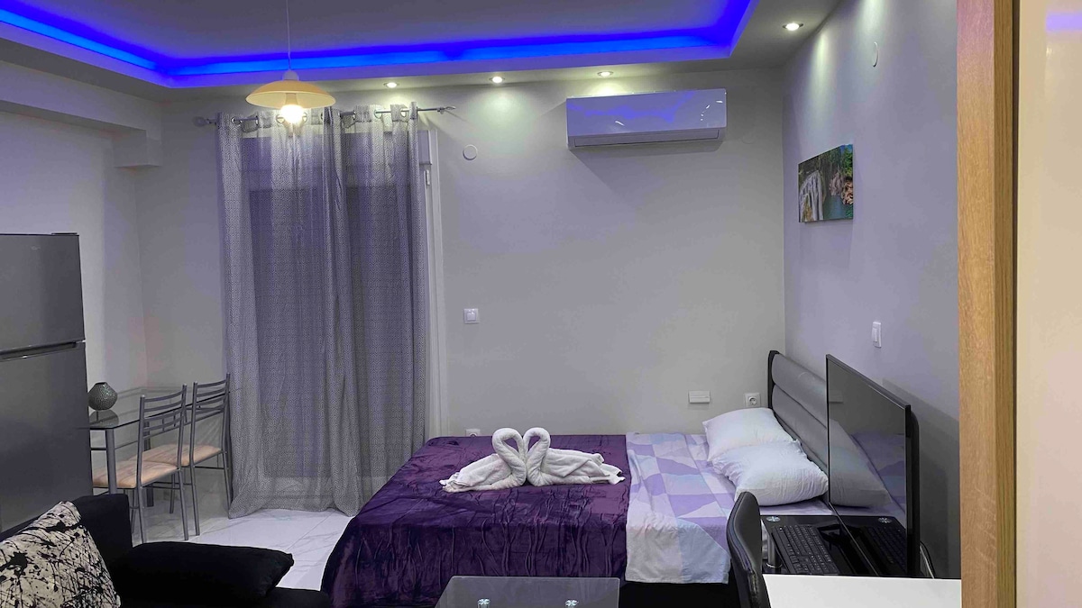 A modern studio space is presented with a double bed covered in a deep purple blanket. A sleek sofa sits adjacent, with a smart TV mounted on the wall. Soft blue lighting highlights the ceiling, while a dining area with metal chairs is visible in the background.