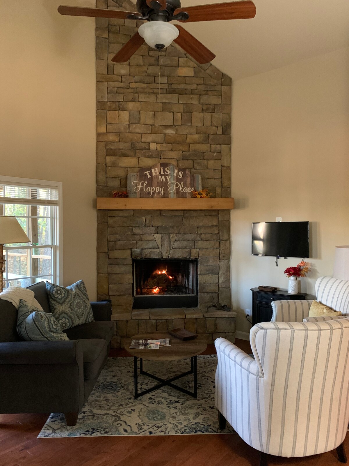 A stone fireplace serves as the focal point of a cozy den, with a comfortable armchair and a dark sofa placed nearby. A coffee table rests on a decorative area rug, and a flat-screen TV is mounted on the wall. Natural light enters through a window beside the fireplace.