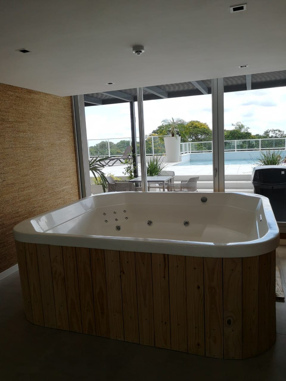 A spacious jacuzzi is positioned against a feature wall with natural textures. Large glass doors provide views of an outdoor area with greenery and a swimming pool. Natural light fills the space, enhancing the welcoming ambiance.