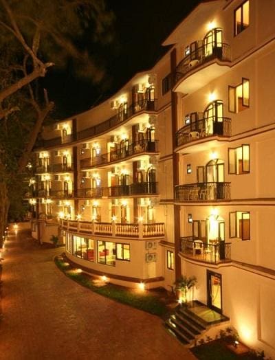 The exterior of the resort is highlighted at night, featuring multiple balconies adorned with soft lighting. The structured facade showcases a mix of windows and doors, with a landscaped pathway leading towards the entrance, enhancing the inviting ambiance.