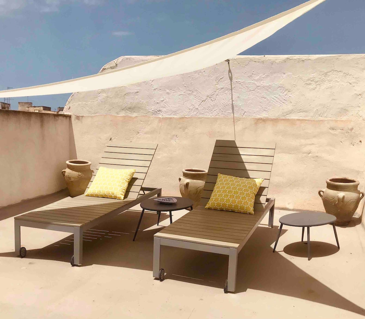 A rooftop terrace is equipped with two loungers, each featuring a patterned yellow pillow. A small round table is positioned nearby, complemented by traditional Sicilian vases. A fabric shade structures protection against direct sunlight.
