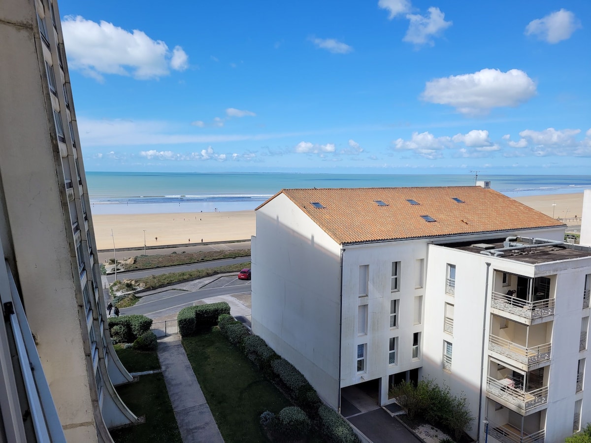 A view of the coastline is displayed, featuring the sandy beach meeting the calm sea under a clear blue sky. The beach is visible in the distance, while neatly arranged buildings and paths are seen in the foreground.