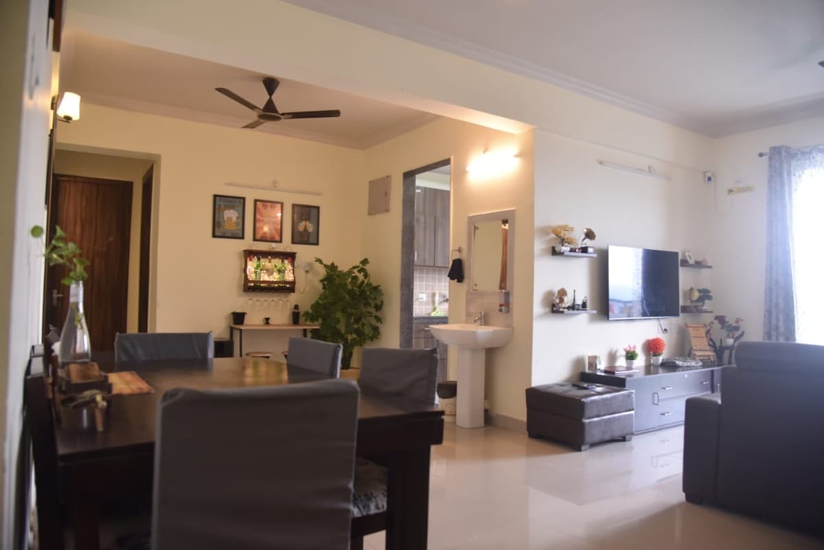 The living and dining area features a spacious table with six chairs, alongside a seating arrangement with dark upholstered furniture. A television is mounted on the wall opposite the sofa, and decorative shelves with plants and art contribute to the inviting ambiance. Soft lighting enhances the overall comfort.